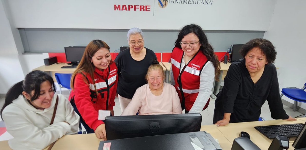 Seis mujeres, entre ellas varias voluntarias con chaleco rojo de MAPFRE, acompañan a una mujer mayor frente a un ordenador en un aula de informática, bajo los logotipos de MAPFRE y Universidad Panamericana.