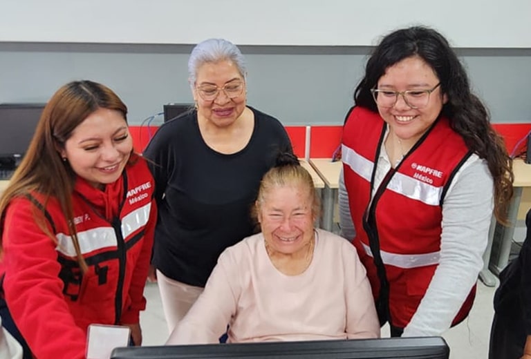 Seis mujeres, entre ellas varias voluntarias con chaleco rojo de MAPFRE, acompañan a una mujer mayor frente a un ordenador en un aula de informática, bajo los logotipos de MAPFRE y Universidad Panamericana.