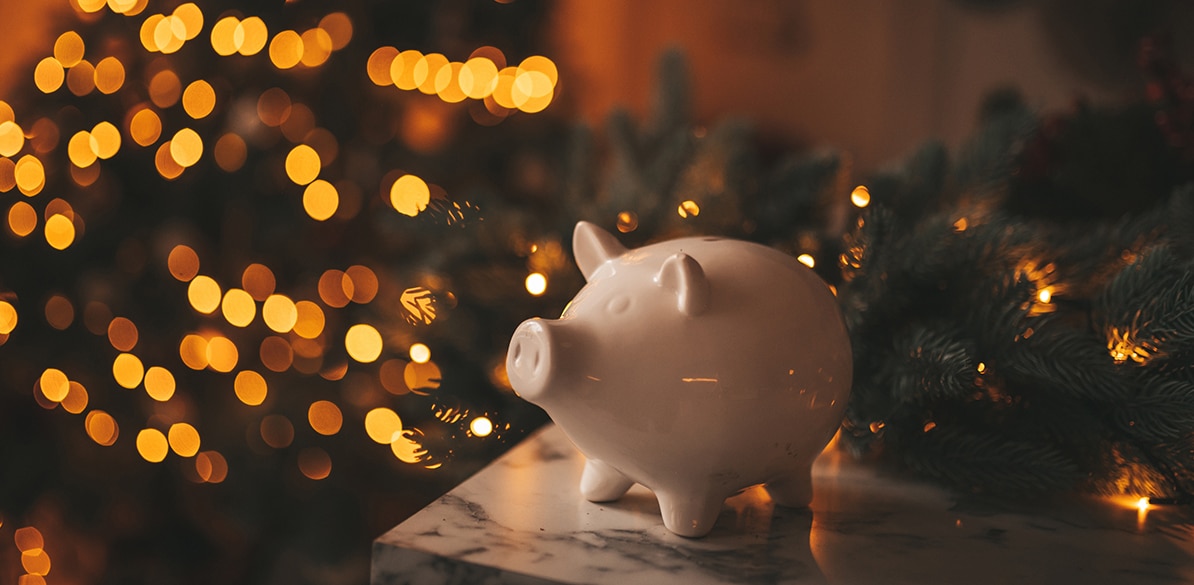 Al frente, una alcancía blanca en forma de cerdito sobre una superficie de mármol; detrás, luces cálidas y un árbol de Navidad desenfocado evocan ahorro y metas financieras navideñas para 2025.
