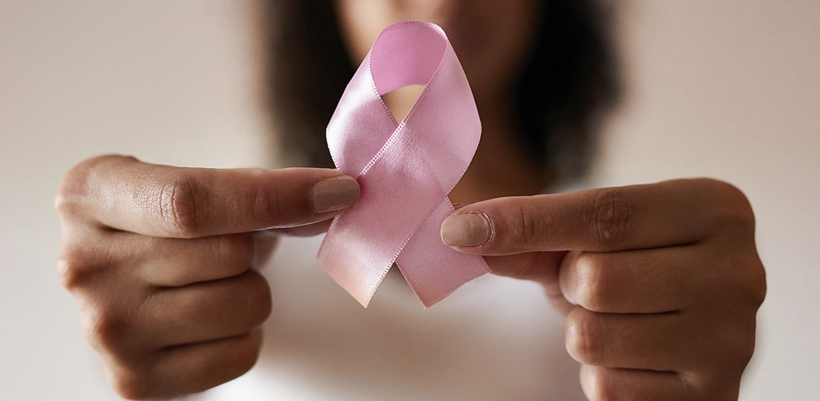 Día Mundial del Cáncer de Mama Primer plano de una mano sosteniendo un lazo rosa, símbolo de la lucha contra el cáncer de mama, en un fondo neutro. La persona está mostrando el lazo con los dedos índice y pulgar de manera destacada, con una ligera profundidad de campo que desenfoca el fondo. El lazo rosa es brillante y está perfectamente formado, destacando sobre la piel de la mano, lo que refuerza el mensaje de conciencia sobre la prevención del cáncer de mama.