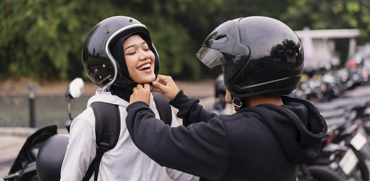 Hacia una movilidad que proteja la vida Una persona con casco negro ayuda a otra a abrochar su casco abierto negro antes de montar en moto, ambas sonriendo en un estacionamiento con más motocicletas al fondo, representando el uso responsable del casco como medida de seguridad vial.