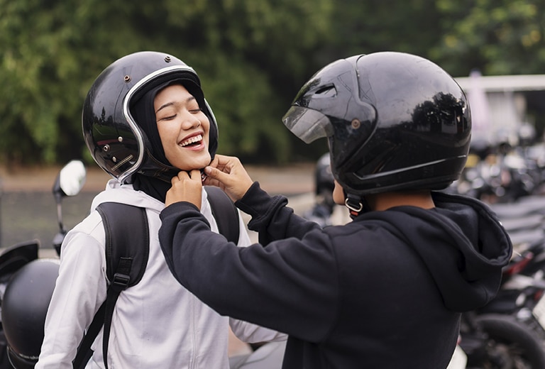 Una persona con casco negro ayuda a otra a abrochar su casco abierto negro antes de montar en moto, ambas sonriendo en un estacionamiento con más motocicletas al fondo, representando el uso responsable del casco como medida de seguridad vial.