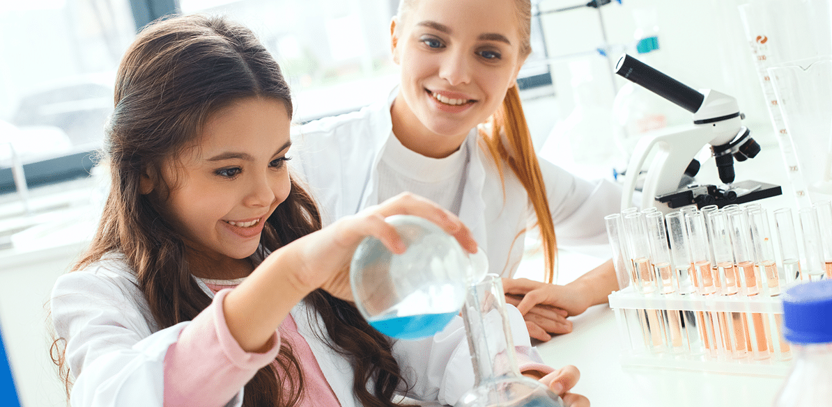 Niña con bata blanca vertiendo líquido azul desde un matraz a una probeta en un laboratorio, junto a una joven mentora; al fondo hay un microscopio y una gradilla con tubos de ensayo con líquidos anaranjados.