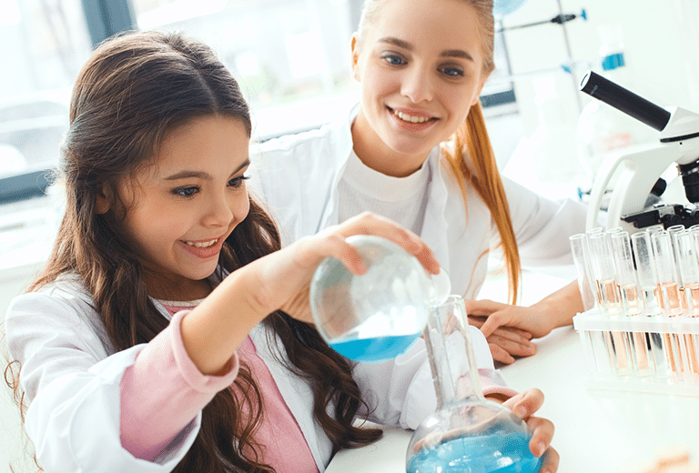 Niña con bata blanca vertiendo líquido azul desde un matraz a una probeta en un laboratorio, junto a una joven mentora; al fondo hay un microscopio y una gradilla con tubos de ensayo con líquidos anaranjados.