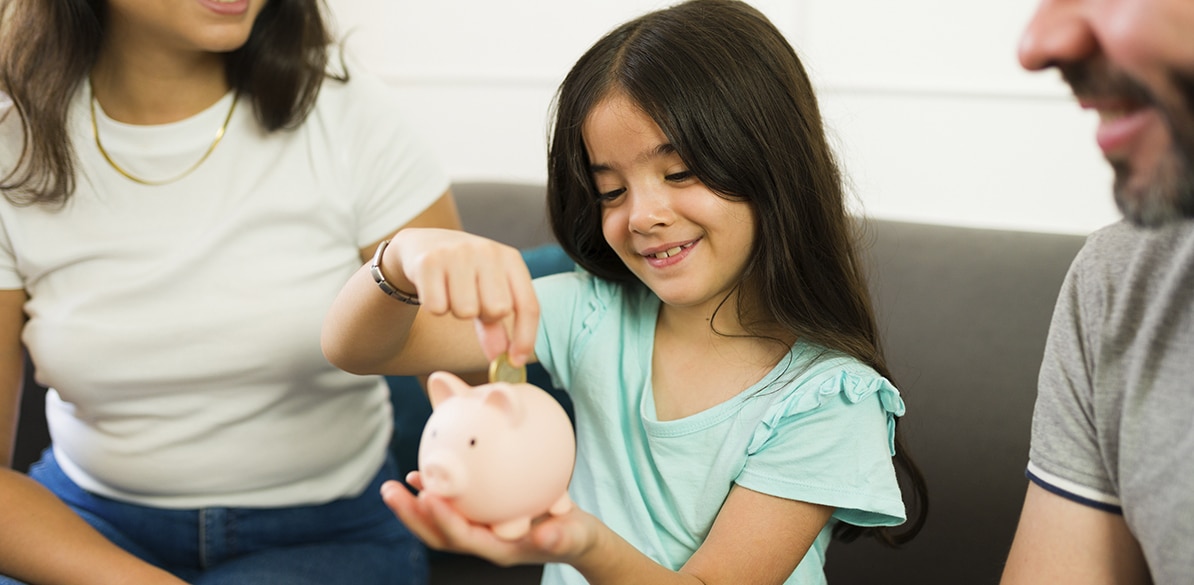 Niña introduce una moneda en una hucha con forma de cerdito mientras sus padres la acompañan, en una escena familiar sobre ahorro y educación financiera.