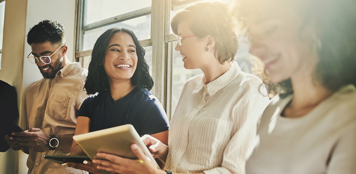 El futuro de México está en manos de la juventud Grupo de cuatro jóvenes sentados junto a una ventana con luz natural, sonriendo y conversando mientras usan una tablet y teléfonos móviles.