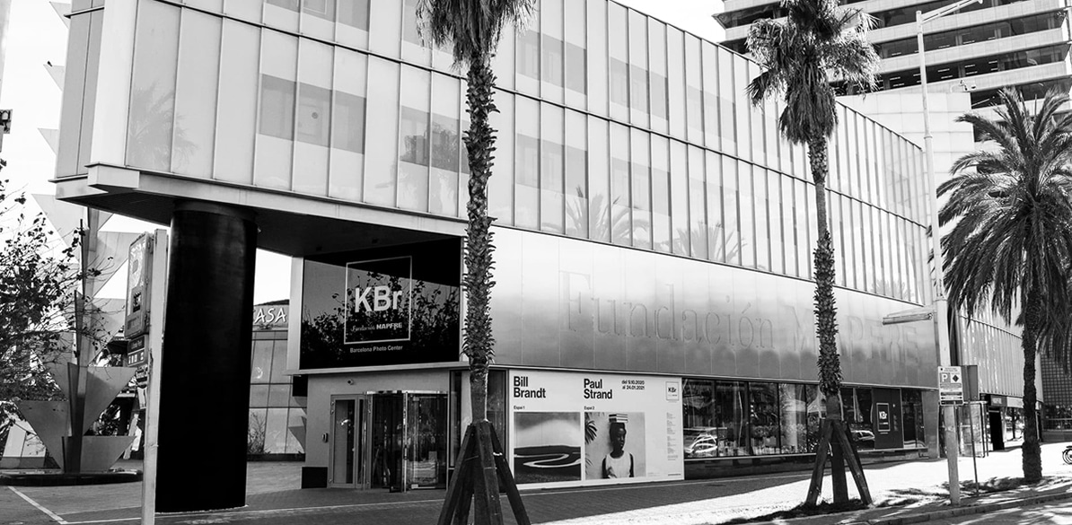 KBr Photo Award Foto en blanco y negro del edificio de la sala de exposiciones fotográficas KBr en Barcelona, con carteles de exposiciones anteriores, de Paul Strand y Bill Brandt, en el centro donde se albergan distintos proyectos fotográficos como los que se darán a conocer en la III Convocatoria del KBr Award.