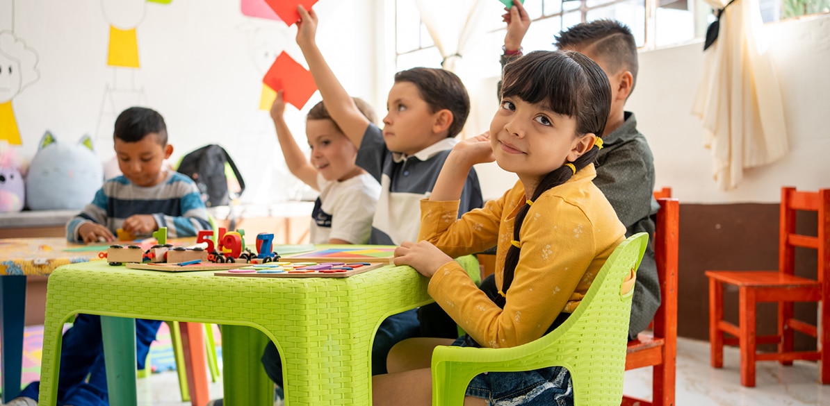 Niña de primaria sentada en una silla verde en un aula, sonriendo a cámara mientras otros niños levantan tarjetas de colores y juegan con materiales educativos sobre una mesa, simbolizando aprendizaje y oportunidades.