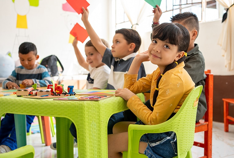 Niña de primaria sentada en una silla verde en un aula, sonriendo a cámara mientras otros niños levantan tarjetas de colores y juegan con materiales educativos sobre una mesa, simbolizando aprendizaje y oportunidades.