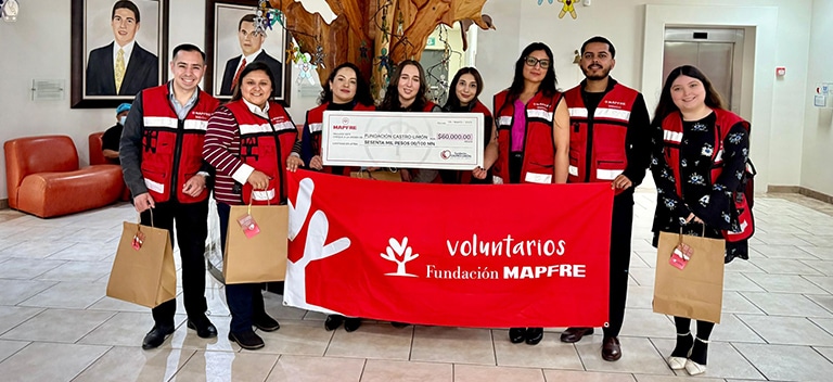 Grupo de voluntarios con chalecos rojos de Fundación MAPFRE posando en interior, sosteniendo una pancarta de Voluntarios Fundación MAPFRE y un cheque, algunos con bolsas en la mano.