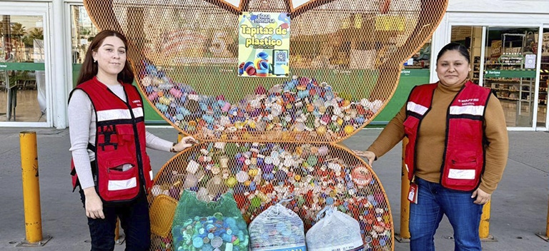 Dos voluntarias con chalecos rojos de Fundación MAPFRE junto a un contenedor en forma de corazón lleno de tapones de plástico, con un cartel de “Tapitas de plástico” visible en la parte superior.