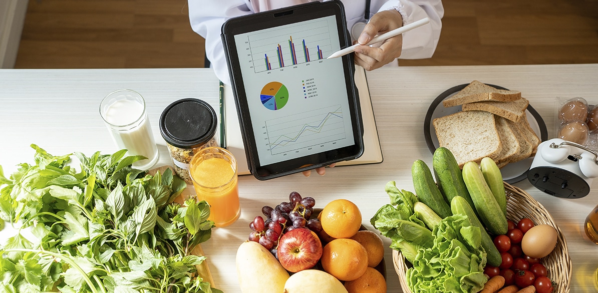 Profesional sanitario señalando con un lápiz óptico una tablet con gráficos de salud y nutrición sobre una mesa con frutas, verduras, pan, huevos, leche y zumo.